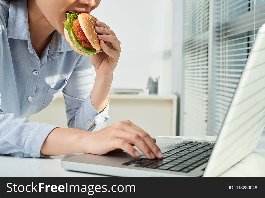 Female entreprepreneur eating chicken burger when working on laptop in office. Female entreprepreneur eating chicken burger when working on laptop in office