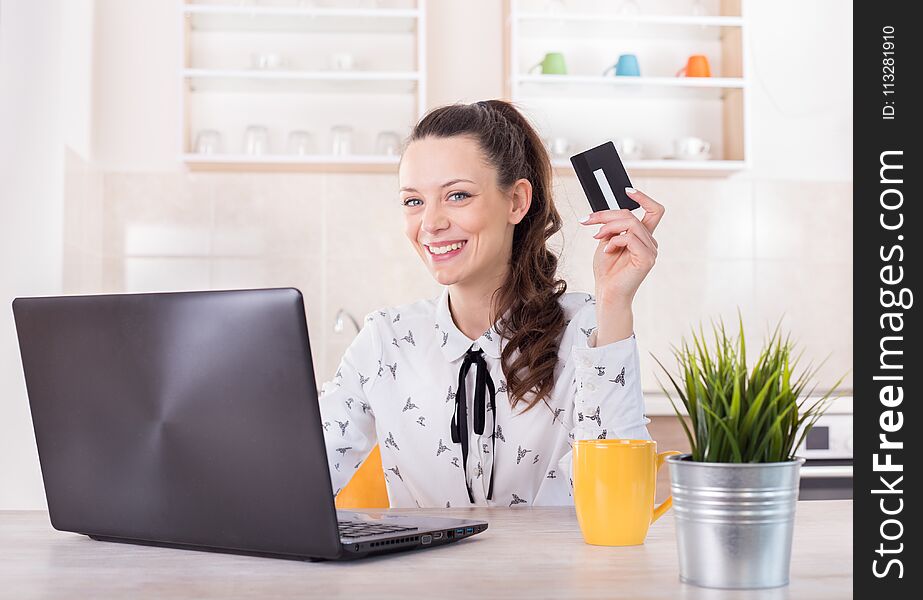 Happy young woman sitting at kitchen table with laptop and holding credit card. Online shopping and e-business concept. Happy young woman sitting at kitchen table with laptop and holding credit card. Online shopping and e-business concept