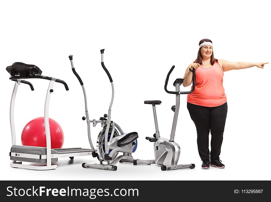 Full length portrait of an overweight woman standing by exercise machines and pointing isolated on white background