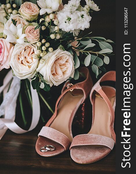 Wedding velvet pink shoes with gold wedding rings beside a bouquet of white roses, eucalyptus on a dark wooden background. Vertical