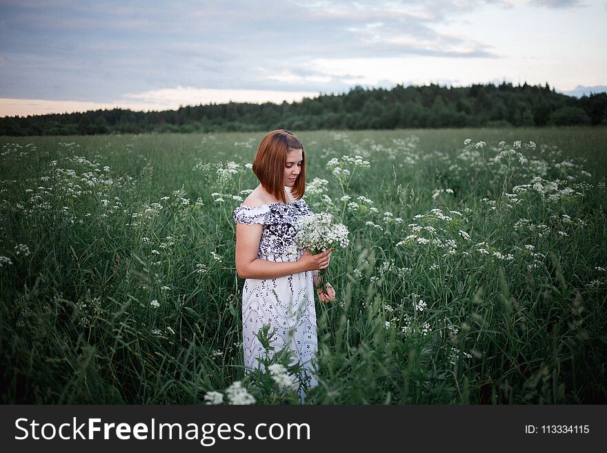 Beautiful brunette in the evening summer field with bouquet of meadow flowers.