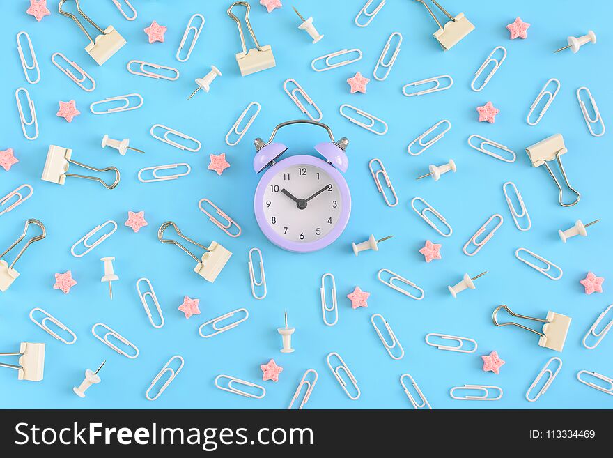 Stationery chaotically scattered on a blue background. White paper clips, clerical buttons and small pink stars in disorder . In the center is a small lilac alarm clock. Beautiful picture from the top.