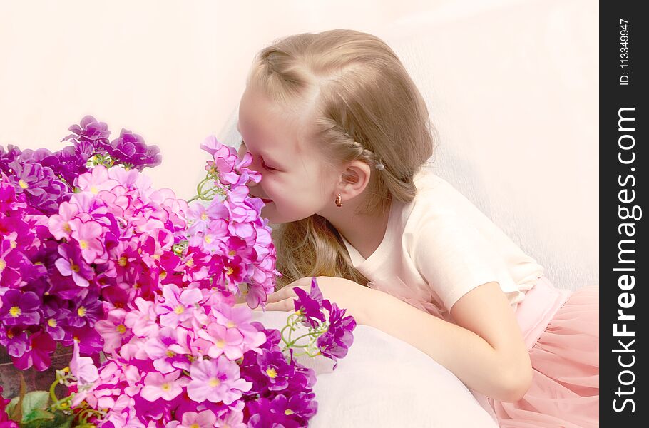 Caucasian little girl with long blond hair, in a beautiful pink skirt and white T-shirt and a pink bow on her chest.Sits on a blue sofa.A bouquet of pink flowers. Caucasian little girl with long blond hair, in a beautiful pink skirt and white T-shirt and a pink bow on her chest.Sits on a blue sofa.A bouquet of pink flowers.