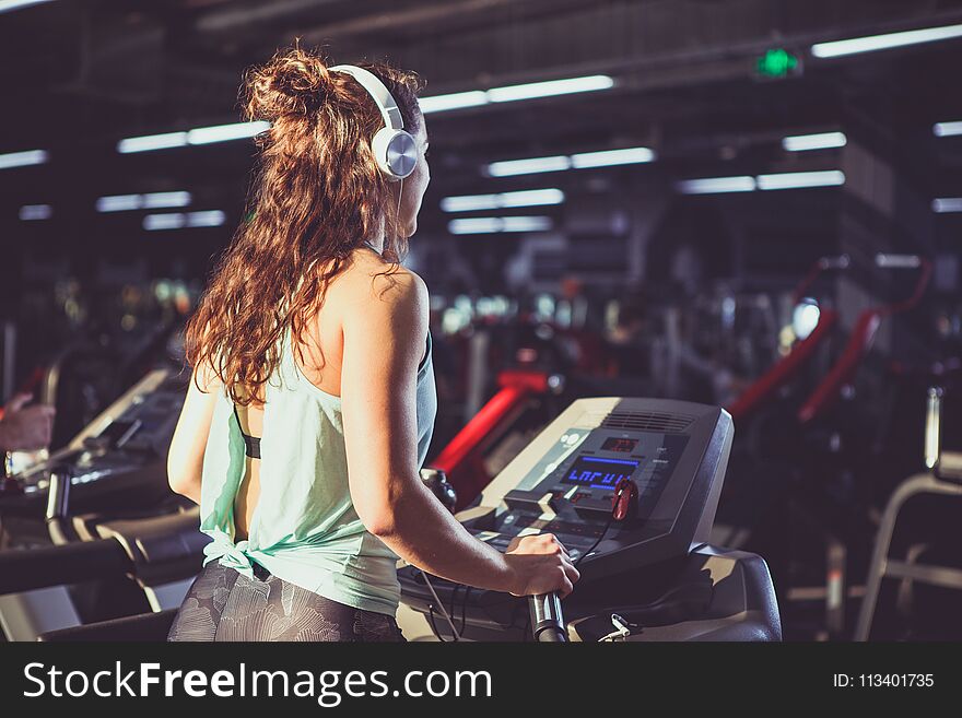 Back view. beautiful woman with lush curly hair in vest and elk runs in gym on treadmill, cardio training machine and weight loss. On head big white headphones, listening music. Sunny weather.