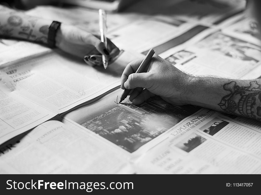 Grayscale Photo of Hands Writing on the Newspaper