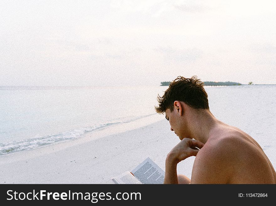 Topless Man Reading Book While Seating at Beach