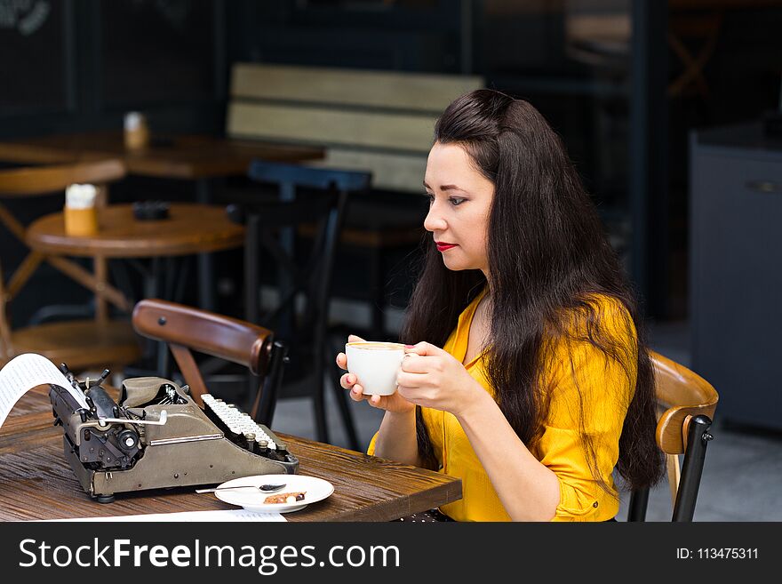 Freelance and writing concept. Writer female with dark long hair, wearing yellow shirt and working in a coffee shop using a retro typewriter. Freelance and writing concept. Writer female with dark long hair, wearing yellow shirt and working in a coffee shop using a retro typewriter.
