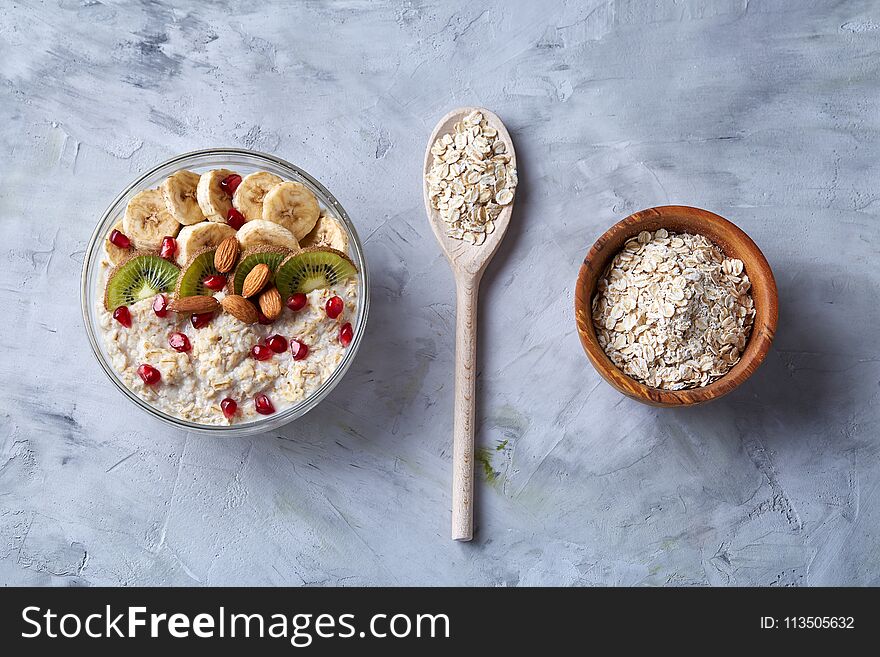 Diet breakfast oatmeal cereal with kiwi, banana, almond and pomergranate seeds and wooden bowl and spoon with oat flakes over white textured background, selective focus, close-up, top view. Healthy eating concept. Breakfast background. Diet breakfast oatmeal cereal with kiwi, banana, almond and pomergranate seeds and wooden bowl and spoon with oat flakes over white textured background, selective focus, close-up, top view. Healthy eating concept. Breakfast background.