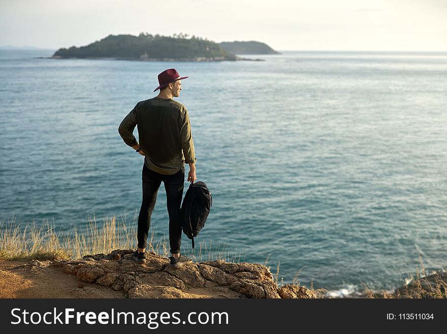 Handsome man with a black backpack stands on the cliff on the background of the green islands in the sea. He wears dark jeans and sneakers, olive shirt and crimson hat. Shoot from the back. Handsome man with a black backpack stands on the cliff on the background of the green islands in the sea. He wears dark jeans and sneakers, olive shirt and crimson hat. Shoot from the back.