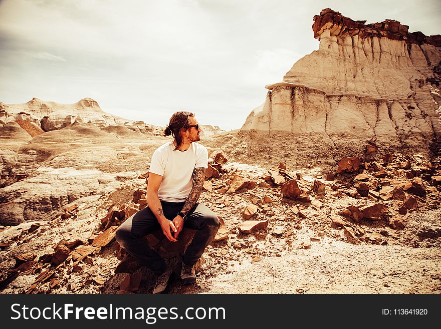Man in White Crew-neck T-shirt and Black Pants Sitting on Boulder Near Cliff