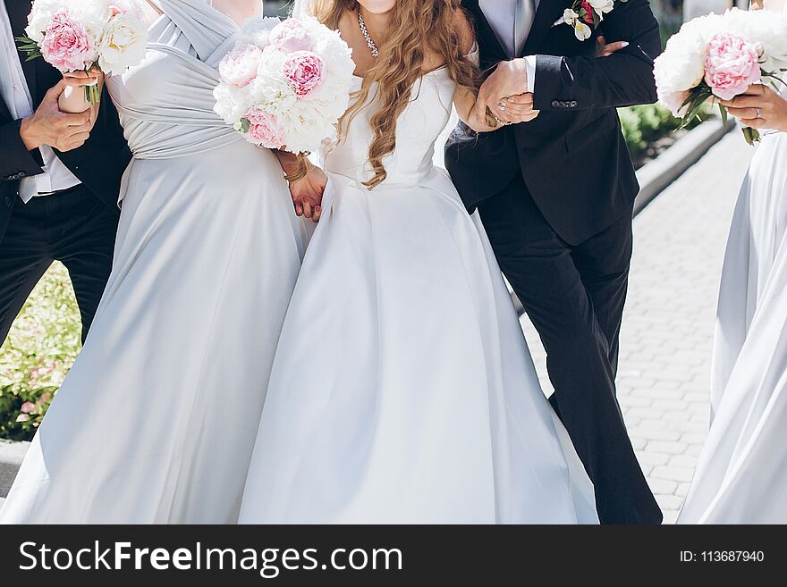 Gorgeous bride with peony bouquet and stylish groom posing in sunny garden with bridesmaids and groomsmen on wedding day. luxury wedding couple having fun with friends