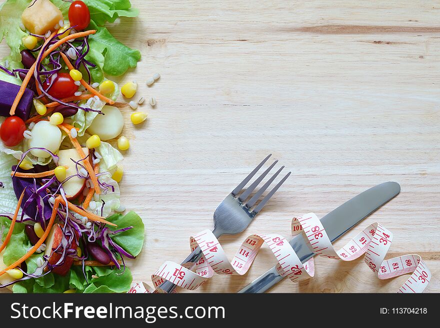 Fresh healthy salad with mixed greens vegetables and fruits on wooden background, with fork, knife, measuring tape, top view with copy space. Healthy balanced diet, weight loss, calorie counting, healthy life and fitness concept.