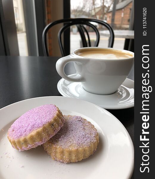 Lavender shortbread cookies on a white plate with a coffee latte in a white mug in the background on a black table in a coffee shop. Lavender shortbread cookies on a white plate with a coffee latte in a white mug in the background on a black table in a coffee shop.