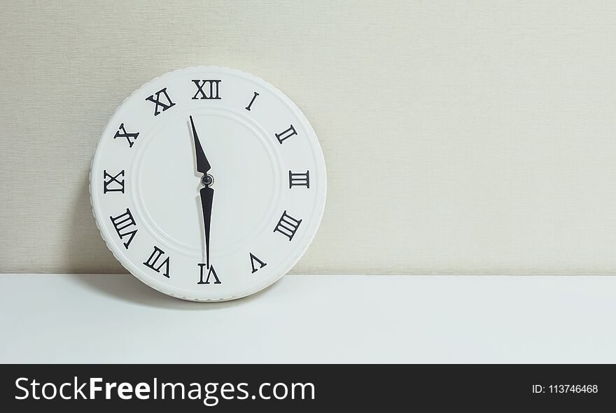 Closeup white clock for decorate show half past eleven or 11:30 a.m. on white wood desk and cream wallpaper textured background wi