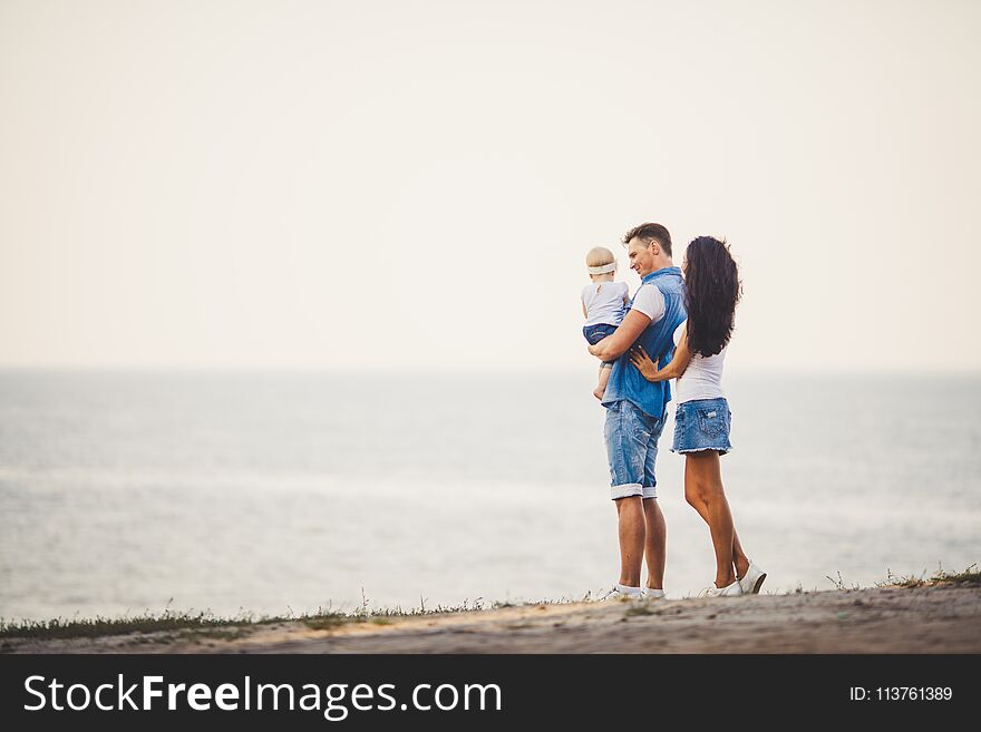 Family holiday in nature. Three, Mom, Dad, daughter one year standing with backs on cliff overlooking sea. man holding hild in arm