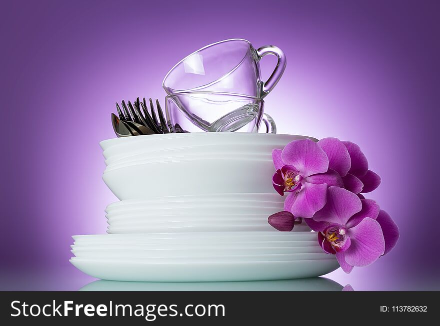 Stack of clean plates, cutlery and cups are top, near Orchid flower on lilac