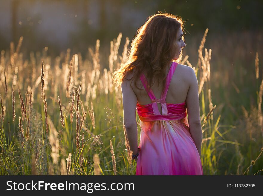 Attractive young woman walking on meadow at sunset - rear view, summer evening