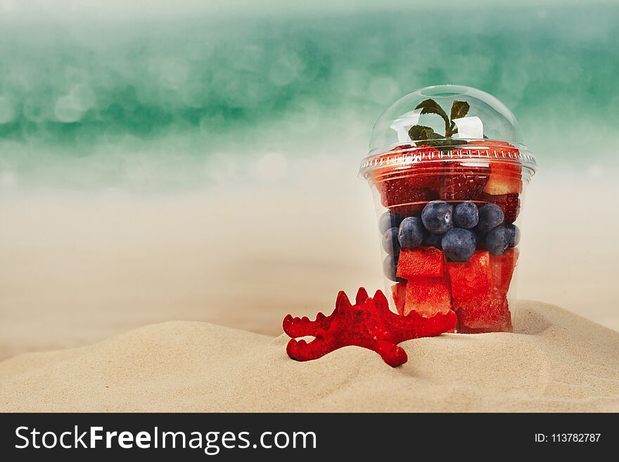 Plastic cup full of fresh cut fruits and berries on the sea coast. Paradise vacations concept. Copy space