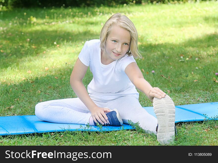 School-age girl in light clothes take sport exercise on a mat in the park. Outdoors workout.