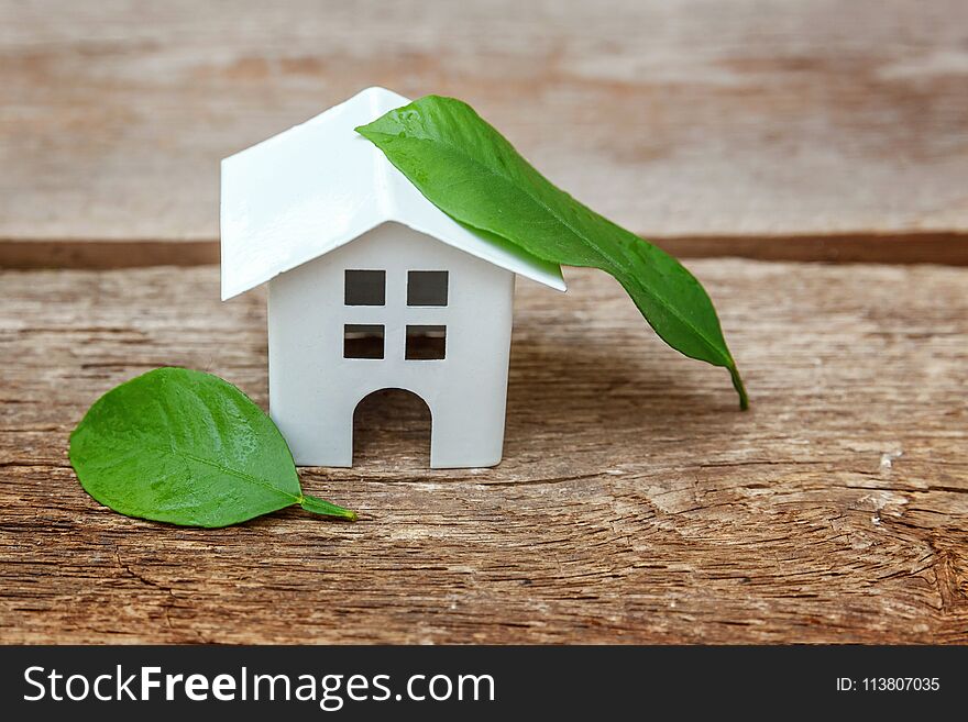 Miniature white toy model house with green leaves on wooden backgdrop. Eco Village, abstract environmental background. Real estate mortgage property insurance dream home ecology concept. Miniature white toy model house with green leaves on wooden backgdrop. Eco Village, abstract environmental background. Real estate mortgage property insurance dream home ecology concept