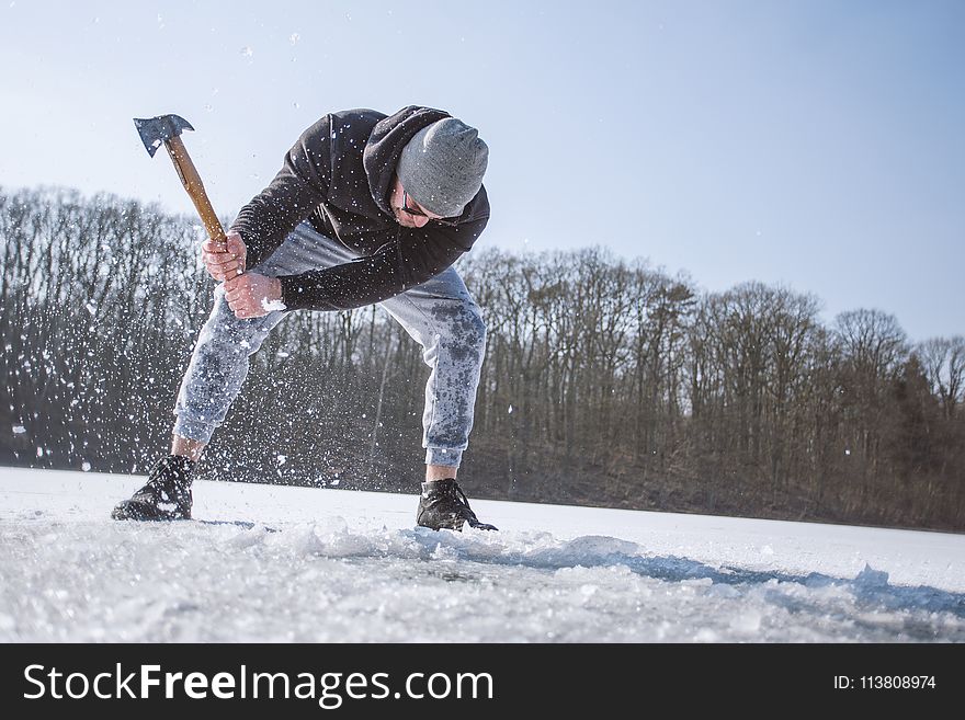 Man Wearing Black Hooded Jacket, Gray Knit Cap, Gray Pants, and Black Shoes Holding Brown Handled Axe While Bending on Snow