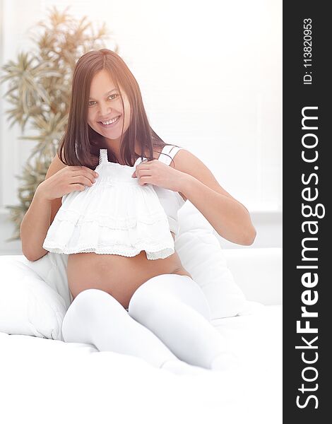Pregnant woman considering children`s shirt , sitting on the bed. the concept of motherhood.