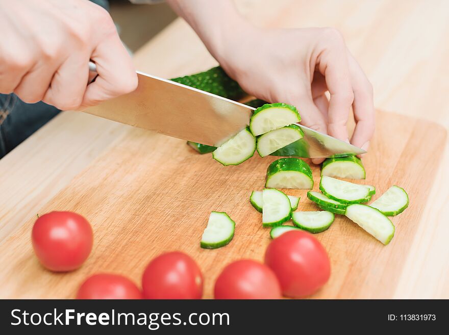 Close-up of female hands cut into fresh cut cucumbers on a wooden cutting board next to pink tomatoes. The concept of