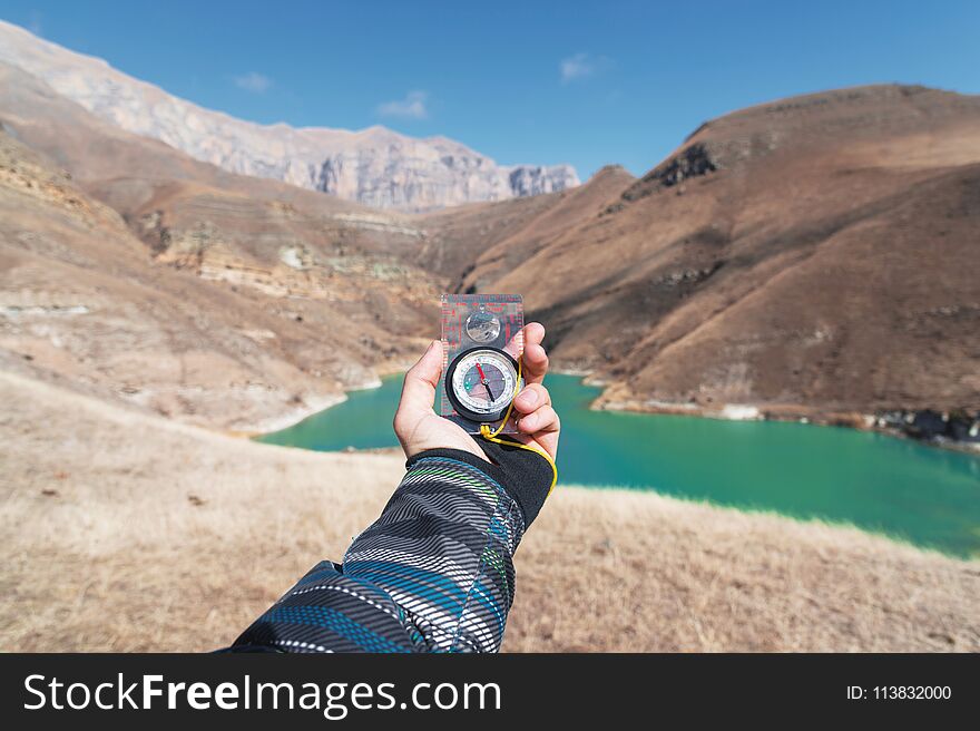 A Man`s Hand Holds A Pocket Magnetic Compass For Navigation Against The Backdrop Of A Rocky Slope And A Mountain Lake