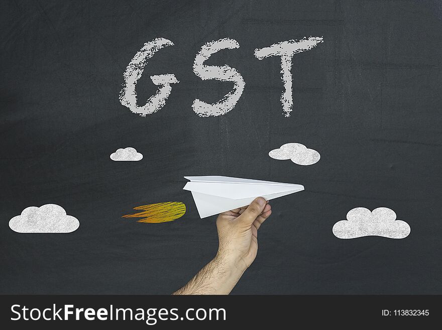 GST Concept. Male Hand Holding Paper Aircraft On Blackboard With Inscription GST