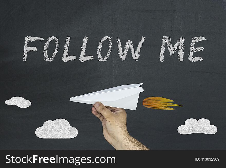 Human hand holding paper plane and Follow me words written on blackboard.