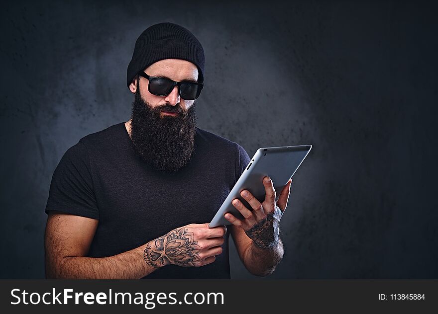 Bearded brutal male in a hat and sunglasses holds tablet PC over grey background. Bearded brutal male in a hat and sunglasses holds tablet PC over grey background.