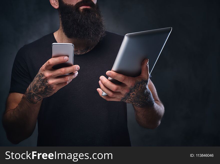 The tattooed, bearded hipster male holds tablet PC and smartphone on grey background. The tattooed, bearded hipster male holds tablet PC and smartphone on grey background.