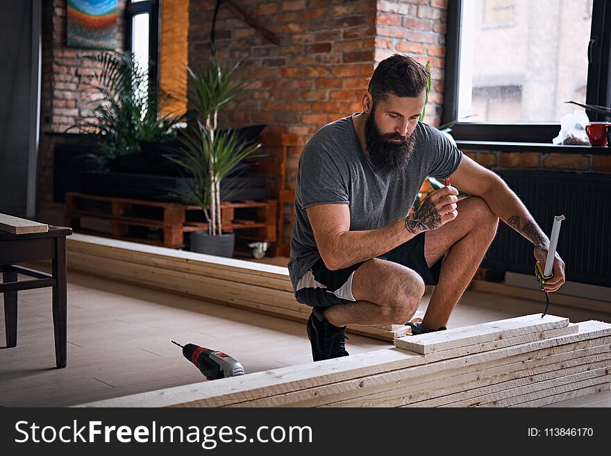 The full body image of bearded tattooed carpenter measuring boards in a room with loft interior.