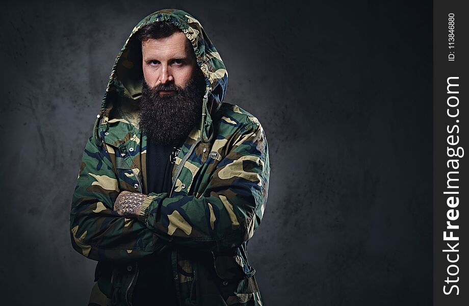 Urban look bearded male with crossed arms dressed in a camouflage jacket on grey background. Urban look bearded male with crossed arms dressed in a camouflage jacket on grey background.