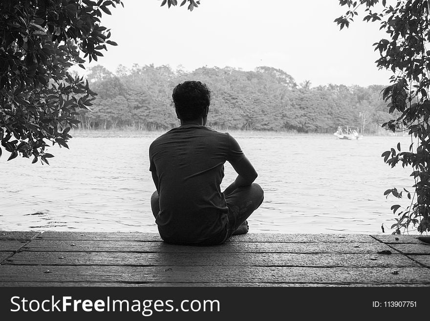 Gray Scale Photography of Man Sitting on Brown Wooden Floor Beside Body of Water