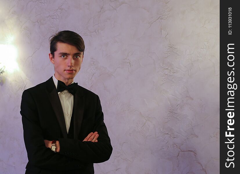 Elegant young guy portrait against light background. Young man in black suit with white shirt and black bow tie. Elegant young guy portrait against light background. Young man in black suit with white shirt and black bow tie.