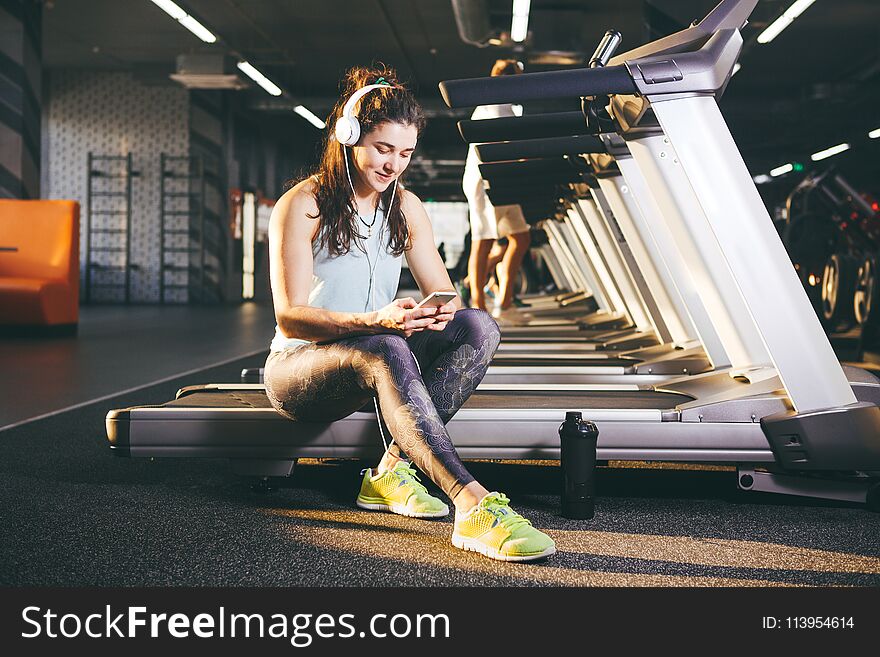 Beautiful young Caucasian girl sportswoman sitting, resting after training on treadmill against the backdrop of gym in sunny weather. Listens to music in sick white headphones, in hand holds the phone