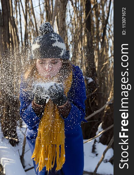 The girl blows off the snow from the hands in the winter forest. She is wearing a purple coat and a gray hat and yellow scarf. The girl blows off the snow from the hands in the winter forest. She is wearing a purple coat and a gray hat and yellow scarf.