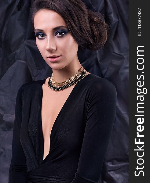 Studio portrait of young beautiful woman in evening dress. Necklace around her neck, hairstyle and makeup. Black dress and dark background