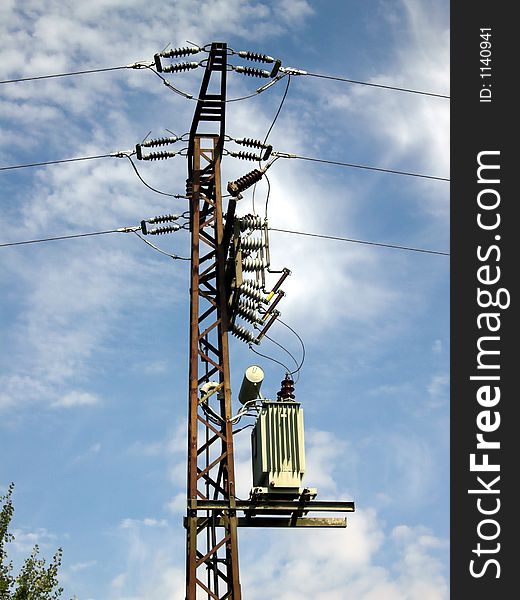 The image shows an old, rusty power pole with its power supply lines. In the background of the picture is a blue sky with some clouds.