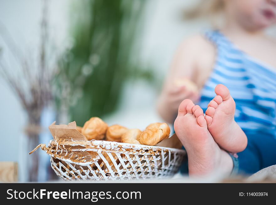 A little girl in a vest with a vest sits on the table. Focus on children`s legs and a basket with croissants
