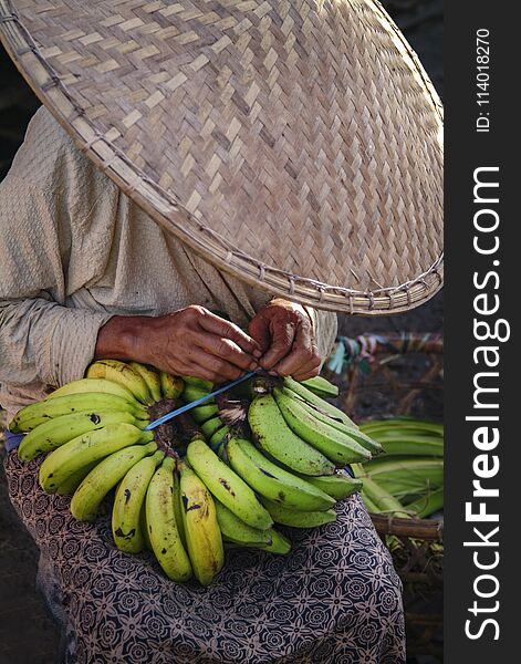 A woman prepares bananas for sale at a traditional public market. She is wearing a conical hat and a sarong. A woman prepares bananas for sale at a traditional public market. She is wearing a conical hat and a sarong.