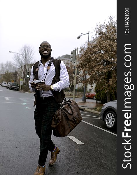 Man In White Button-up Dress Shirt Holding Camera While Smiling And Crossing Street