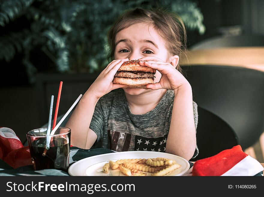 Little girl in T-shirt with American flag in cafe eating a burger and drinking a drink, concept of America Day and American food. Little girl in T-shirt with American flag in cafe eating a burger and drinking a drink, concept of America Day and American food