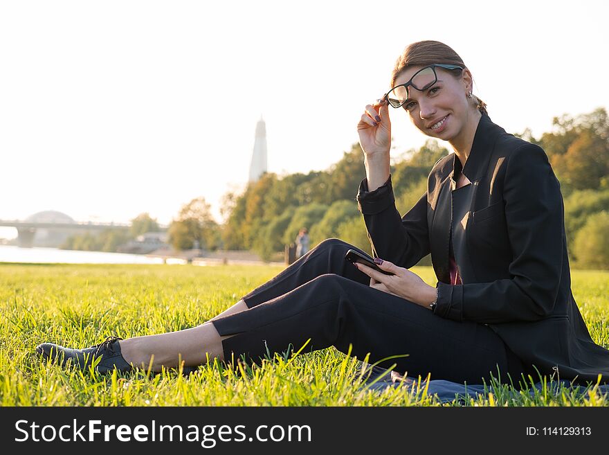 Young businesswoman in glasses using her smartphone and sitting on green grass at sunny day. Working outdoor, staying connected. Young businesswoman in glasses using her smartphone and sitting on green grass at sunny day. Working outdoor, staying connected