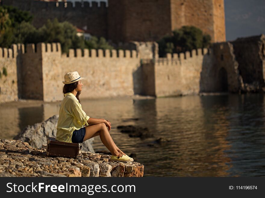 Sideview potrait of a solo traveller woman sitting on vintage suitcase by the sea and wearing dark blue shorts, yellow cotton shirt and hat. Copy space. Sideview potrait of a solo traveller woman sitting on vintage suitcase by the sea and wearing dark blue shorts, yellow cotton shirt and hat. Copy space.