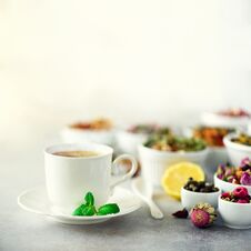 Tea Concept With Copy Space. Different Kinds Of Dry Tea In White Ceramic Bowls And Cup Of Aromatic Tea On Grey Stock Photos