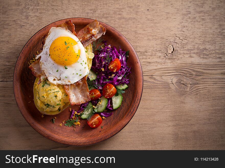 Baked potato in jacket loaded with cheddar cheese and topped with bacon and fried egg in plate on wooden table.