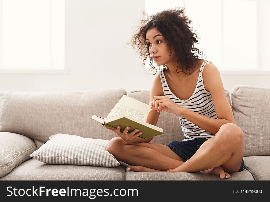 Thoughtful african-american student girl reading book. Young woman studying at home, sitting on beige couch, copy space. Thoughtful african-american student girl reading book. Young woman studying at home, sitting on beige couch, copy space