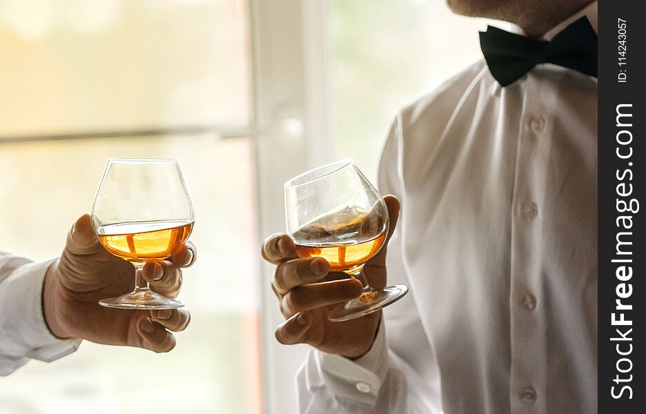 Two glasses of whiskey in the hands of men in white shirts and bow ties. Secular evening, toast, friendship
